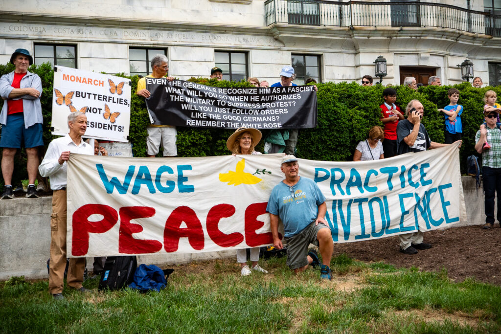 Demonstrators stand along the parade route carrying banners in protest of President Donald Trump and advocating non-violence during the Army 250th Anniversary Parade in Washington, DC Saturday, June 14. Photo by Hannah Parcells.
